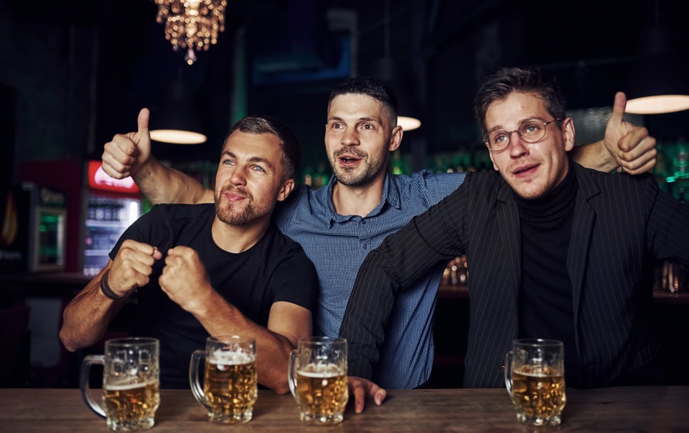 Three men sit together at a bar with arms around each other, smiling and gesturing enthusiastically, with three mugs of beer on the table in front of them. The bar is dimly lit with a chandelier above.