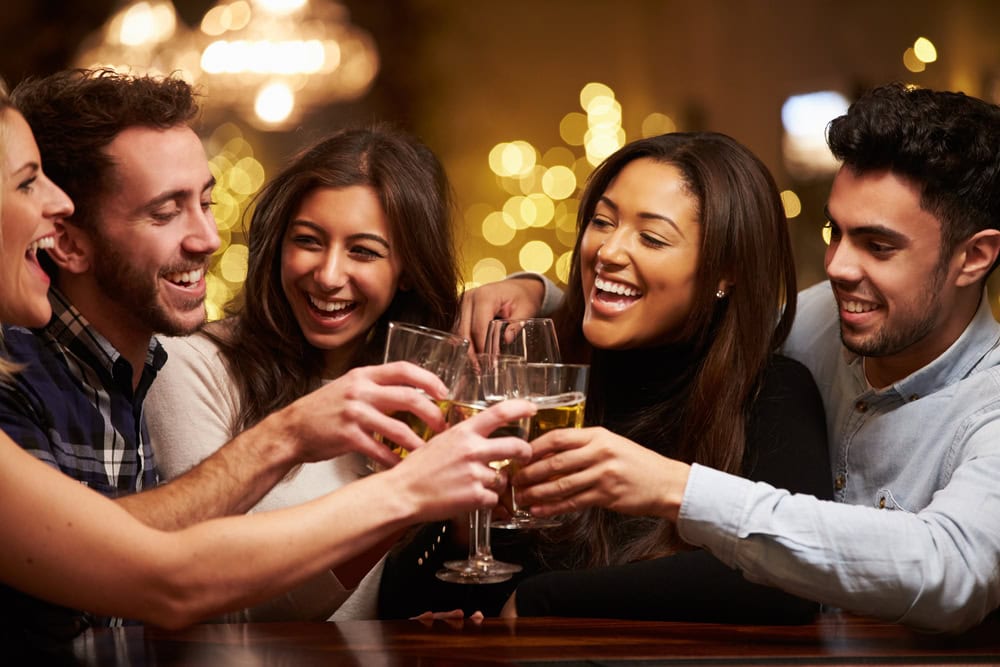 Five friends sit together at a bar, smiling and clinking glasses in a toast. Warm, festive string lights glow in the background, creating a cheerful and celebratory atmosphere.
