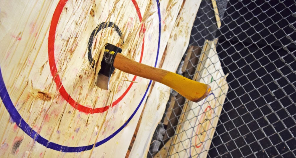 A yellow-handled axe is embedded in the center of a wooden target with red, blue, and black rings; a chain-link fence is visible in the background.